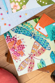 A colourful mosaic art piece featuring two dragonflies decorated with patterned tiles, shown on a wooden surface alongside other craft supplies. Moulin_Roty_-_Les_Rosalies_-_Mosaic_Craft_Kit_-_Strawberries_Cream_-_Ingrid_Lewis_-_Designer
